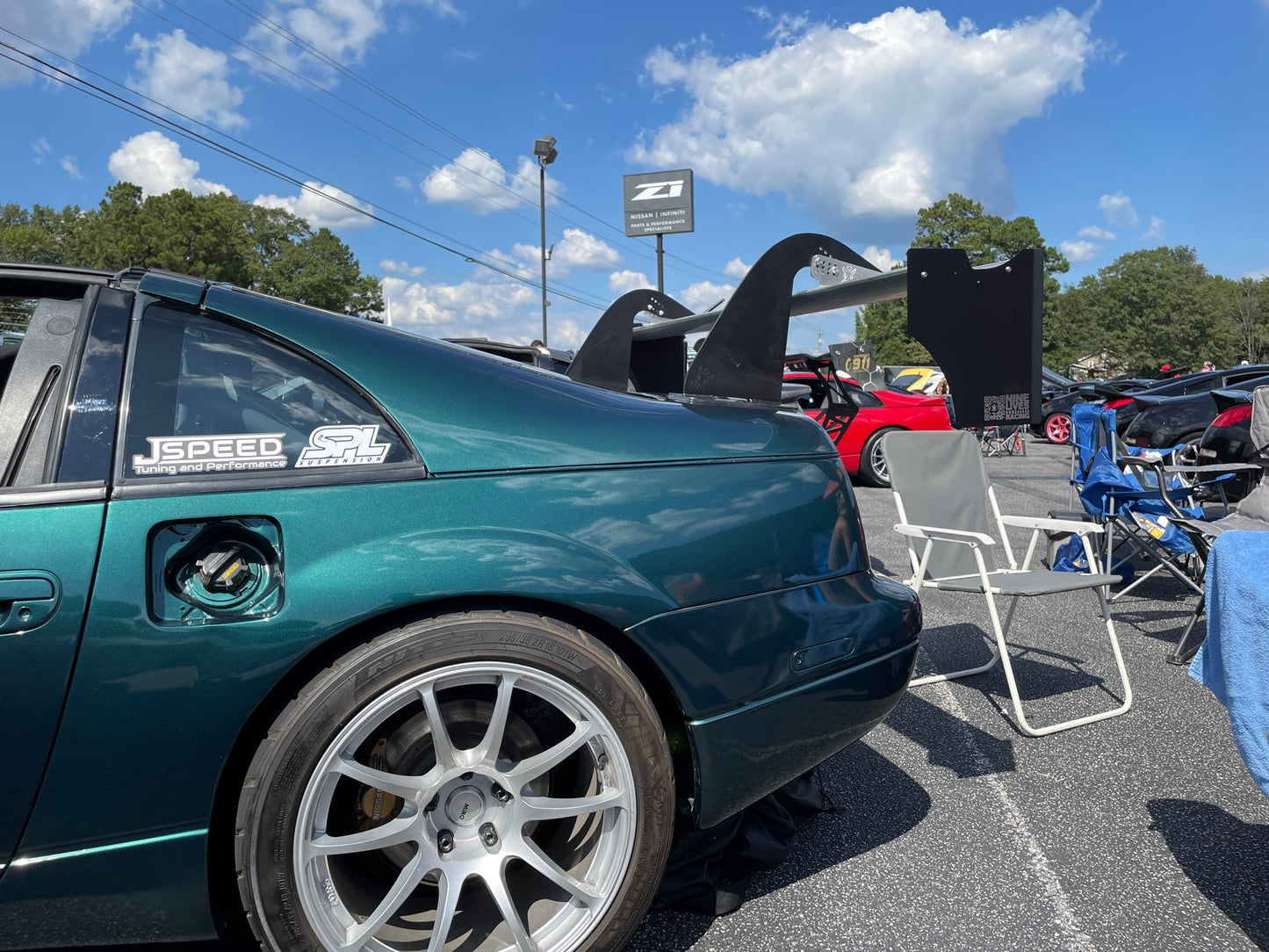 A green sports car with a large rear wing, specifically a 300ZX Big Wang kit '88-'00 Z32 from 9livesracing, is parked at an outdoor car show. The car's gas cap is open, and it has a "JSpeed" sticker on the rear side window. Blue skies and scattered clouds are visible in the background, along with other parked cars and folding chairs.