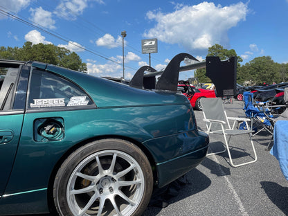 A green sports car with a large rear wing, specifically a 300ZX Big Wang kit '88-'00 Z32 from 9livesracing, is parked at an outdoor car show. The car's gas cap is open, and it has a "JSpeed" sticker on the rear side window. Blue skies and scattered clouds are visible in the background, along with other parked cars and folding chairs.