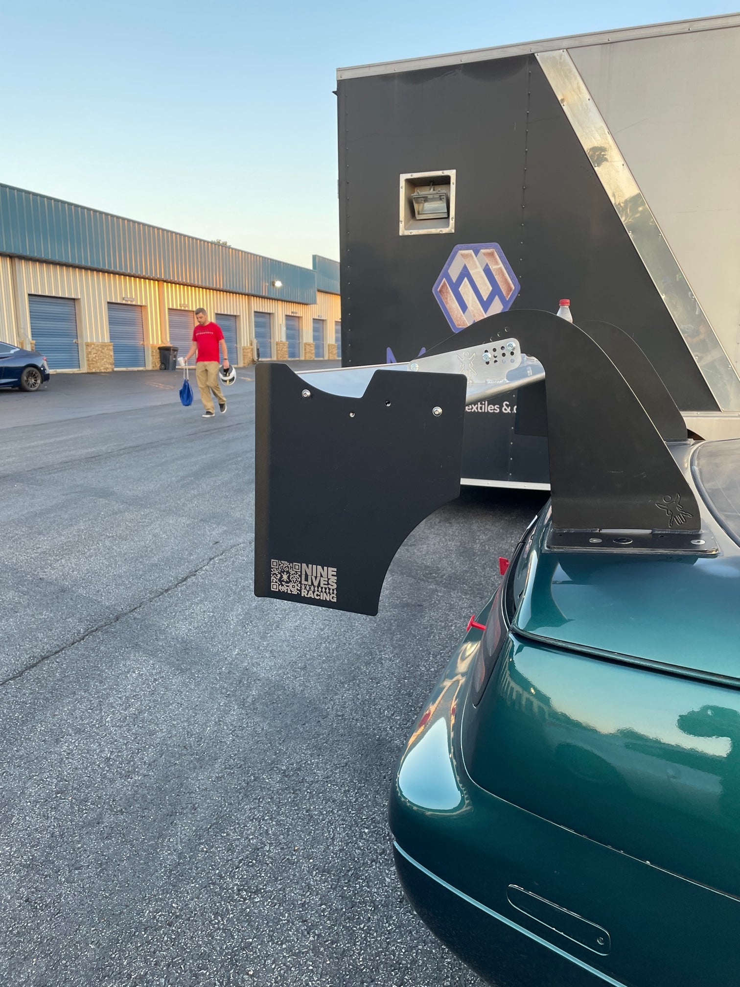 A green sports car is parked outside a row of industrial buildings. The car's large 300ZX Big Wang kit '88-'00 Z32 from 9livesracing bears a QR code and the text "DUNE RACING." This setup optimizes down-force, enhancing its lift-to-drag ratio. In the background, a person in a red shirt walks away, carrying shopping bags.