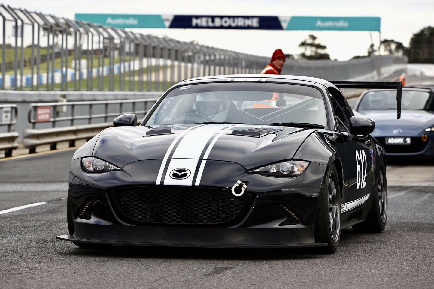 A black Mazda race car with white stripes and the number 842 on the side, equipped with a Miata Big Chassis mount Wang Kit 2016+ ND from 9livesracing for added down-force, is parked on a racetrack near a pit lane during an event in Melbourne, Australia. Barriers line the track, and a person in an orange vest stands in the background.