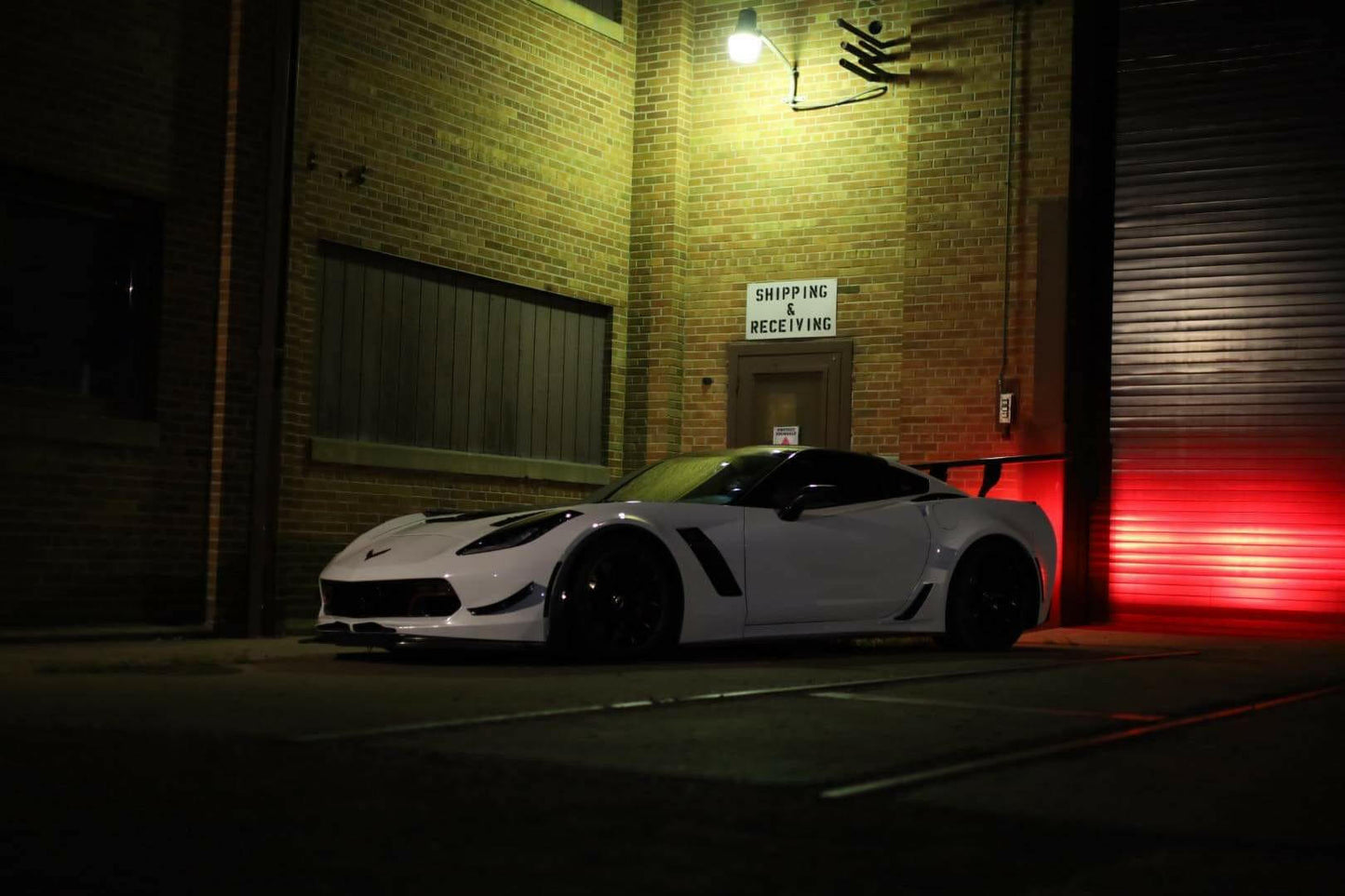 A low-profile, sleek, white sports car fitted with a 9livesracing Corvette Big Wang ZR1 replacement wing C7 is parked outside a brick building under a dim light, near a sign that reads "Shipping and Receiving." The car's shadow is cast on the ground, and red light from the right adds contrast to the scene.