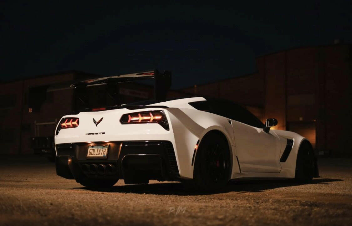 A white sports car with a visible Corvette Big Wang ZR1 replacement wing C7 by 9livesracing and dark-tinted windows is parked in a dimly lit area at night. The car's license plate reads "GOLDZN." Its high down-force design stands out against the industrial-style buildings with minimal lighting.