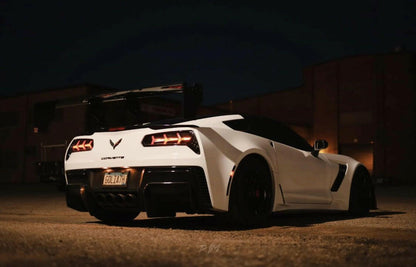 A white sports car with a visible Corvette Big Wang ZR1 replacement wing C7 by 9livesracing and dark-tinted windows is parked in a dimly lit area at night. The car's license plate reads "GOLDZN." Its high down-force design stands out against the industrial-style buildings with minimal lighting.
