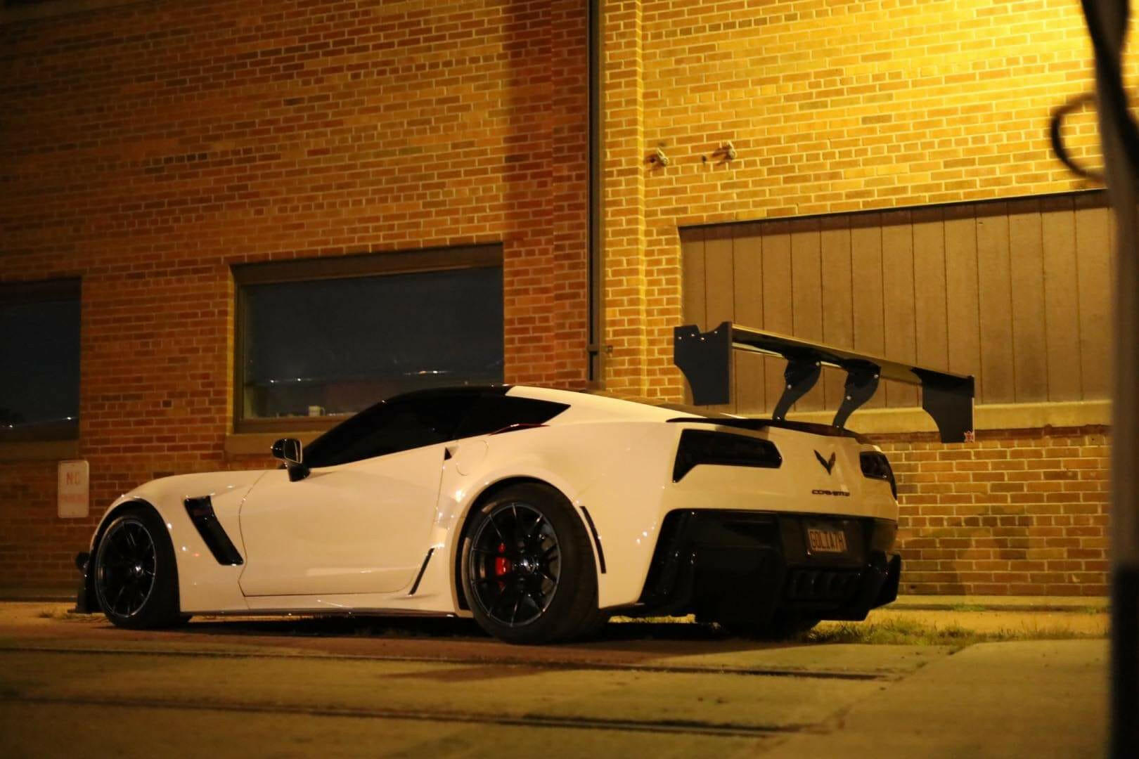 A sleek white sports car with black wheels and a 9livesracing Corvette Big Wang ZR1 replacement wing C7 is parked beside a brick building at night. The building is illuminated by a yellowish streetlight, casting a warm glow on the high down-force rear spoiler.