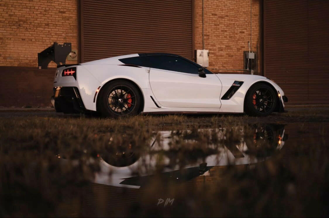 A sleek white sports car, likely a Corvette with the 9livesracing Corvette Big Wang ZR1 replacement wing C7, is parked by a brick industrial building. Its image is reflected in a puddle in the foreground. The setting appears to be during dusk, with soft lighting highlighting the high down-force wings and the car's contours and details.