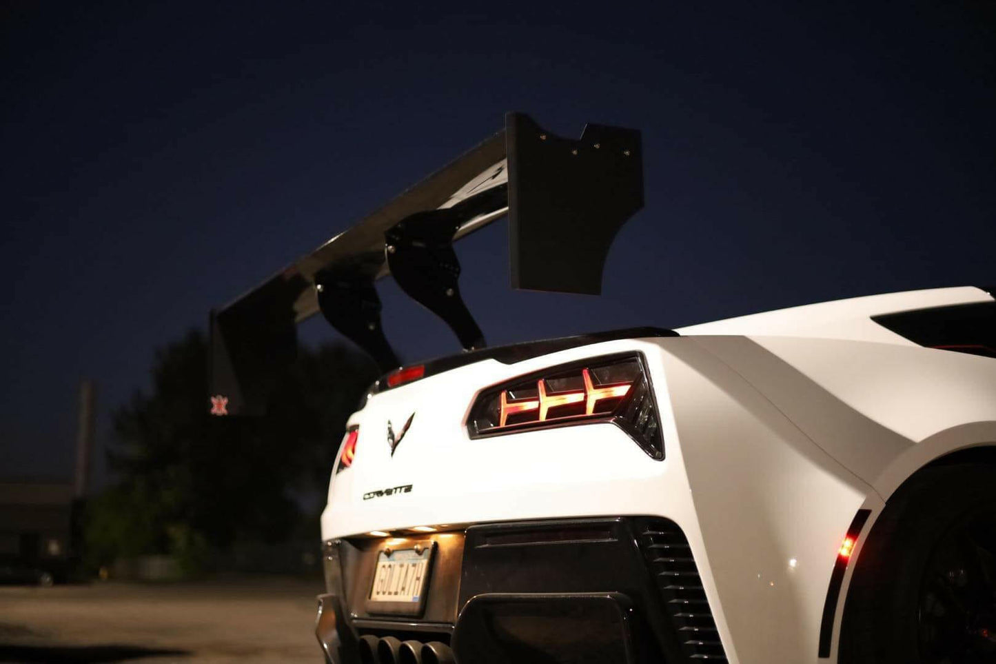 A close-up of the rear end of a white sports car at night, showcasing prominent taillights and a 9livesracing Corvette Big Wang ZR1 replacement wing C7. The car's license plate and surrounding environment are partially visible in the background.