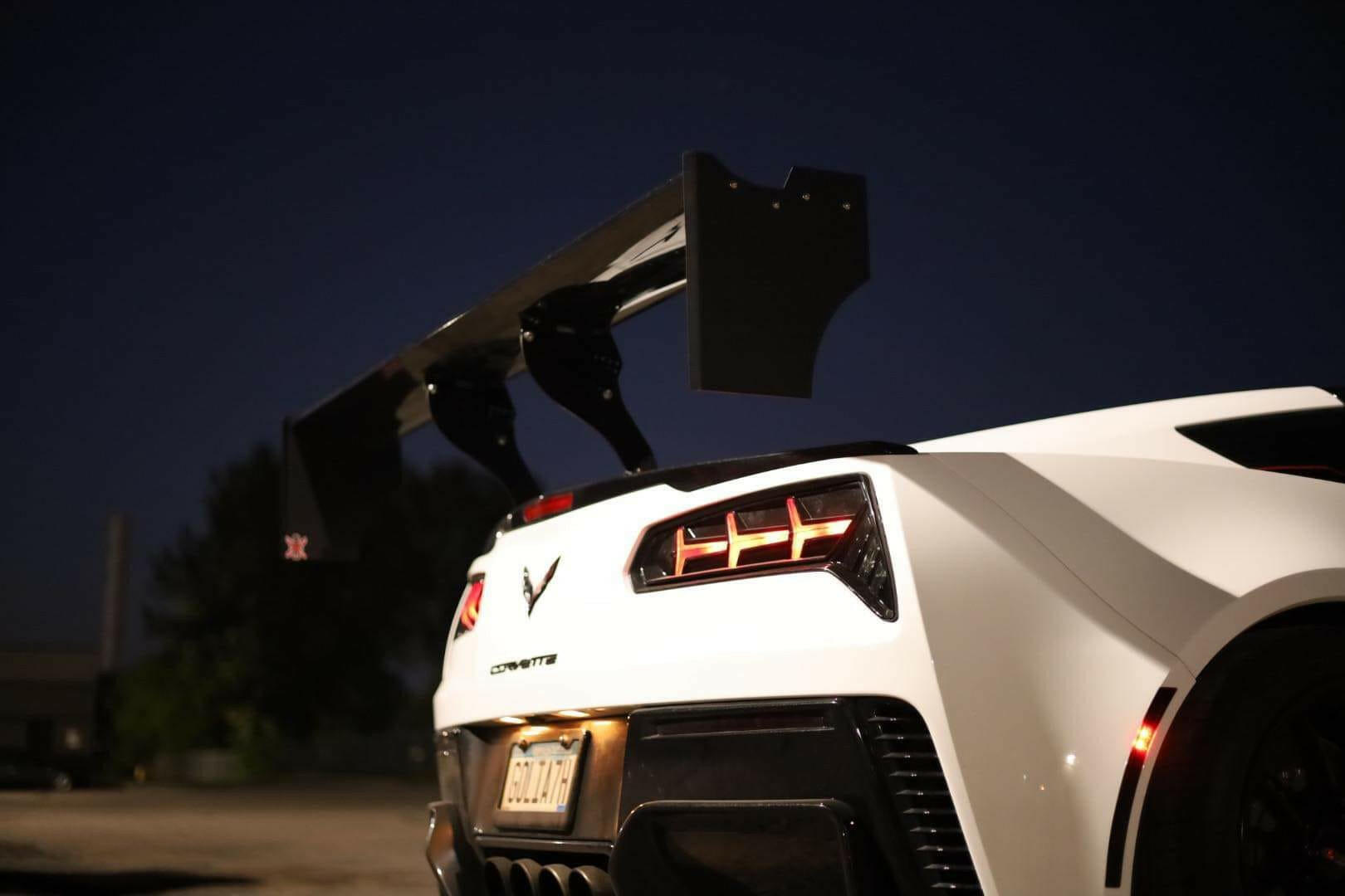 A close-up of the rear end of a white sports car at night, showcasing prominent taillights and a 9livesracing Corvette Big Wang ZR1 replacement wing C7. The car's license plate and surrounding environment are partially visible in the background.