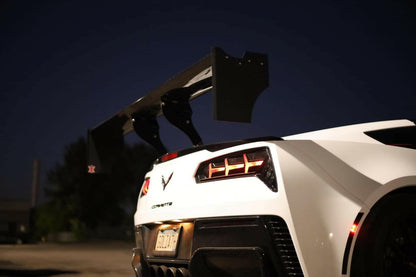 A close-up of the rear end of a white sports car at night, showcasing prominent taillights and a 9livesracing Corvette Big Wang ZR1 replacement wing C7. The car's license plate and surrounding environment are partially visible in the background.