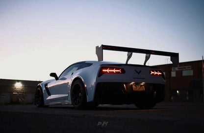In a dimly lit urban area during twilight, a white sports car equipped with the Corvette Big Wang ZR1 replacement wing C7 by 9livesracing is parked. The taillights are illuminated, and the car's low-profile design and sleek body are highlighted by the surrounding shadows and soft evening light. Its high down-force ensures superior road grip.