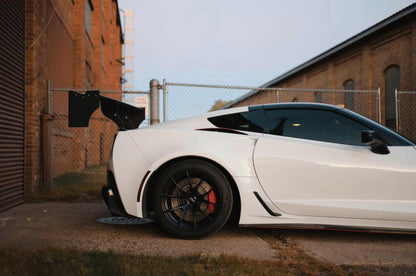 A white sports car with a large 9livesracing Corvette Big Wang ZR1 replacement wing C7 is parked beside a chain-link fence and brick building. The car has black rims and red brake calipers. The extruded aluminum wings add impressive high down-force performance, enhancing its sleek appearance in the industrial setting.