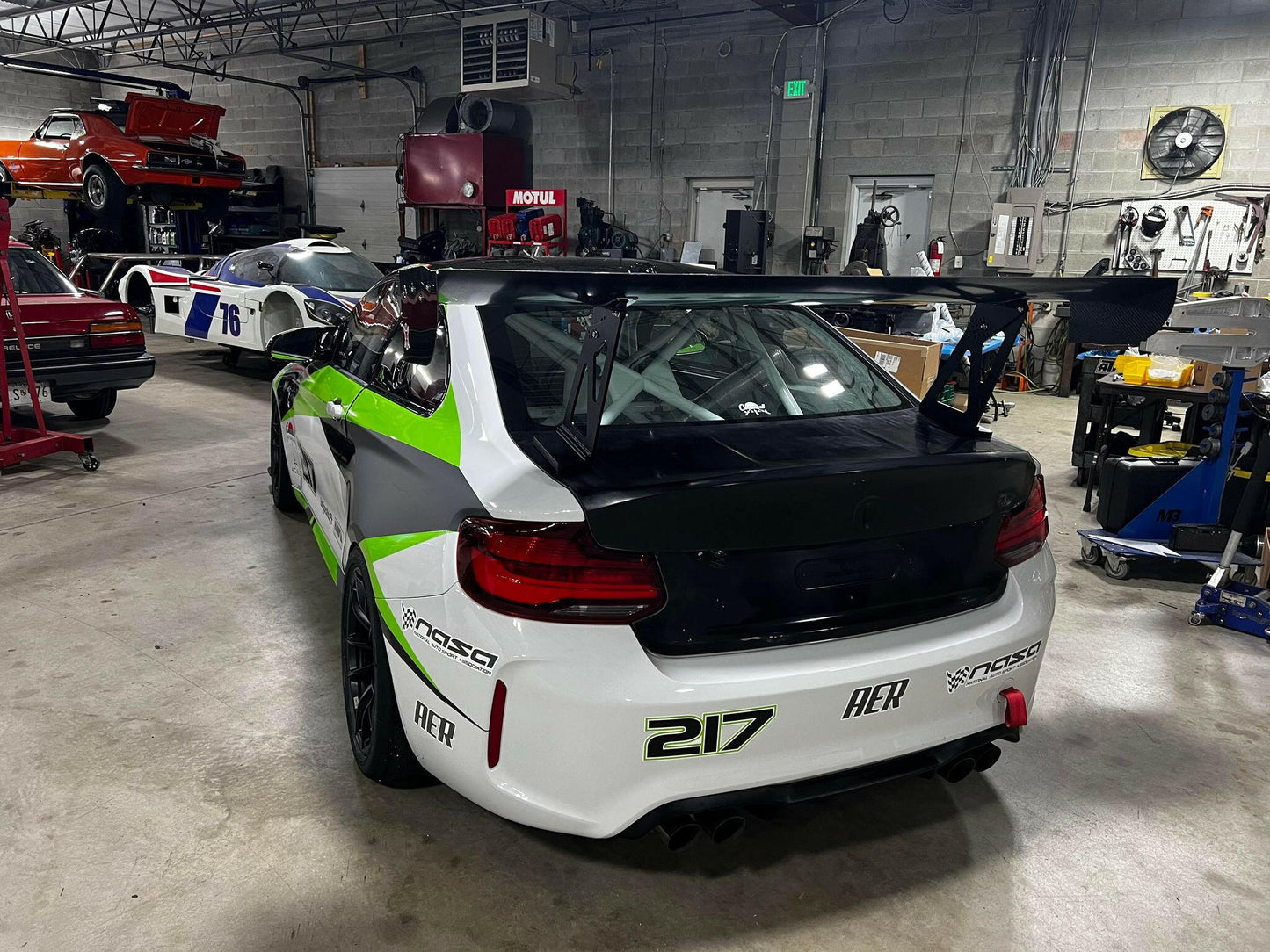 A BMW M2 CS Racing equipped with the 9livesracing Big Wang Kit, sporting green and white livery and numbered 217, is parked in a garage. The car features various sponsor logos. In the background, other vehicles, including a red car and a white car with the number 75, are visible alongside garage equipment.