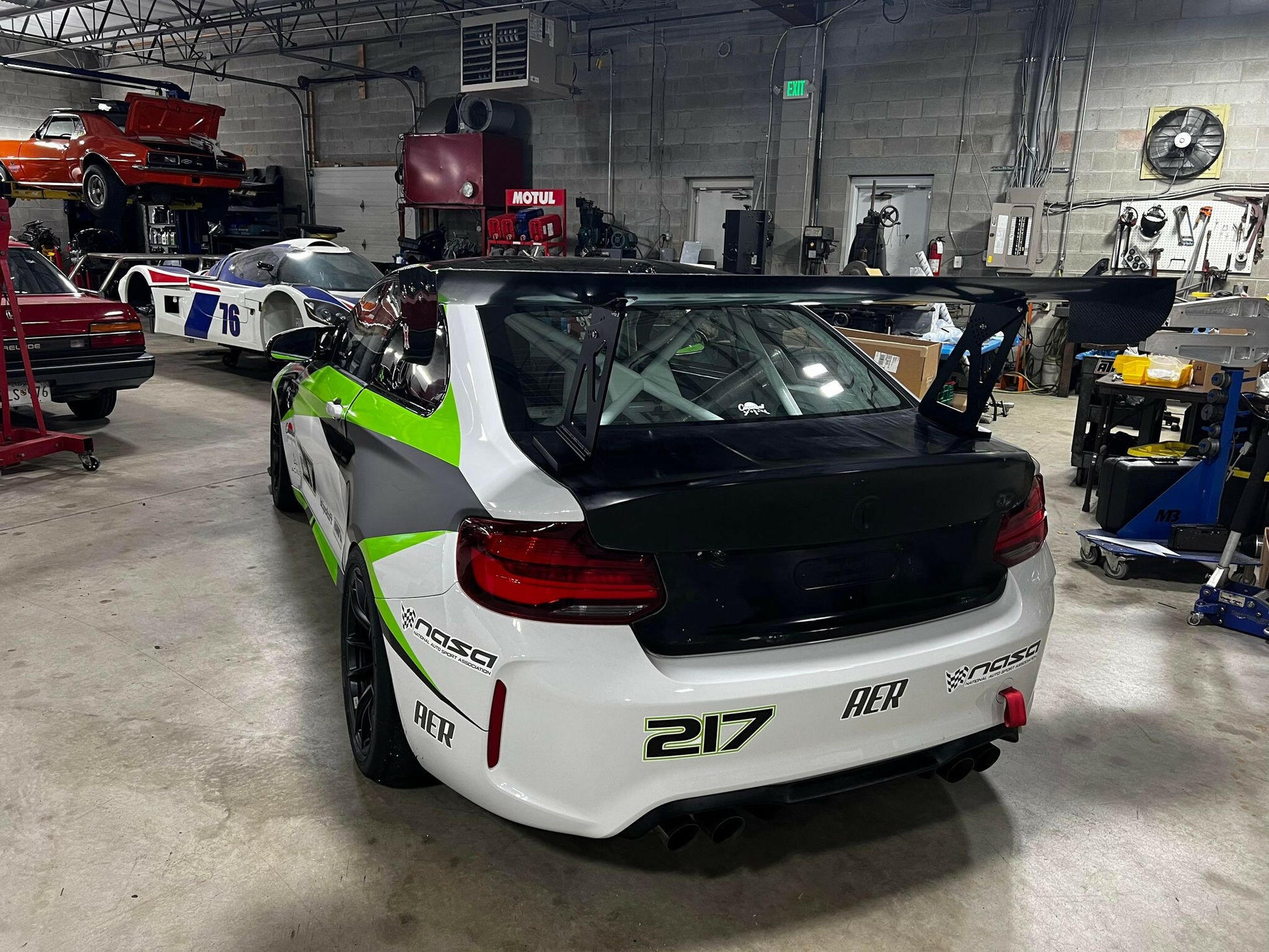 A BMW M2 CS Racing equipped with the 9livesracing Big Wang Kit, sporting green and white livery and numbered 217, is parked in a garage. The car features various sponsor logos. In the background, other vehicles, including a red car and a white car with the number 75, are visible alongside garage equipment.
