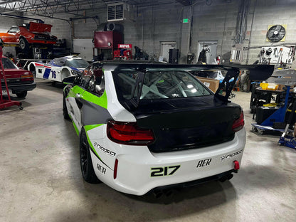 A BMW M2 CS Racing equipped with the 9livesracing Big Wang Kit, sporting green and white livery and numbered 217, is parked in a garage. The car features various sponsor logos. In the background, other vehicles, including a red car and a white car with the number 75, are visible alongside garage equipment.