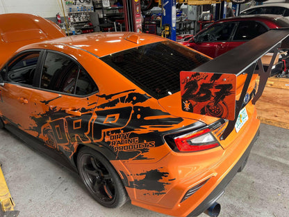 A Subaru Impreza sedan in orange, equipped with the 9livesracing Subaru Impreza STI Big Wang kit '2022 - current. The vehicle features a prominent rear spoiler and proudly displays a "Dirty Racing Products" decal. On the back left panel, there's an intricate black and orange dragon design. Optimized for downforce adjustments, the car is parked in a garage with various tools and equipment visible in the background.