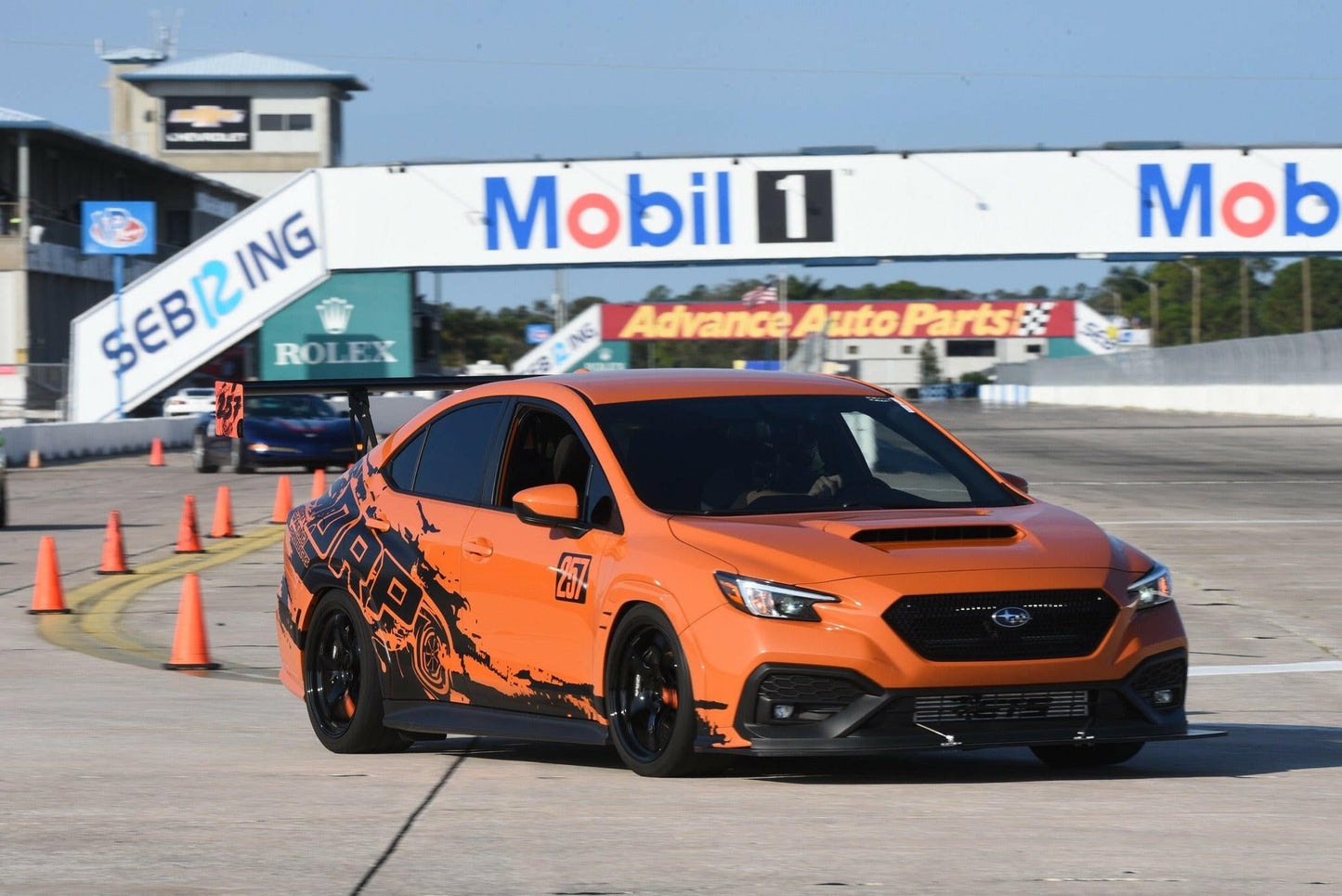 A Subaru Impreza STI Big Wang kit '2022 - current from 9livesracing, featuring an orange and black design with the number 77 on the side and precise downforce adjustments, speeds along the track at Sebring International Raceway. Various sponsor banners, including Mobil 1 and Advance Auto Parts, are visible in the background. Orange safety cones line the edges of the track.