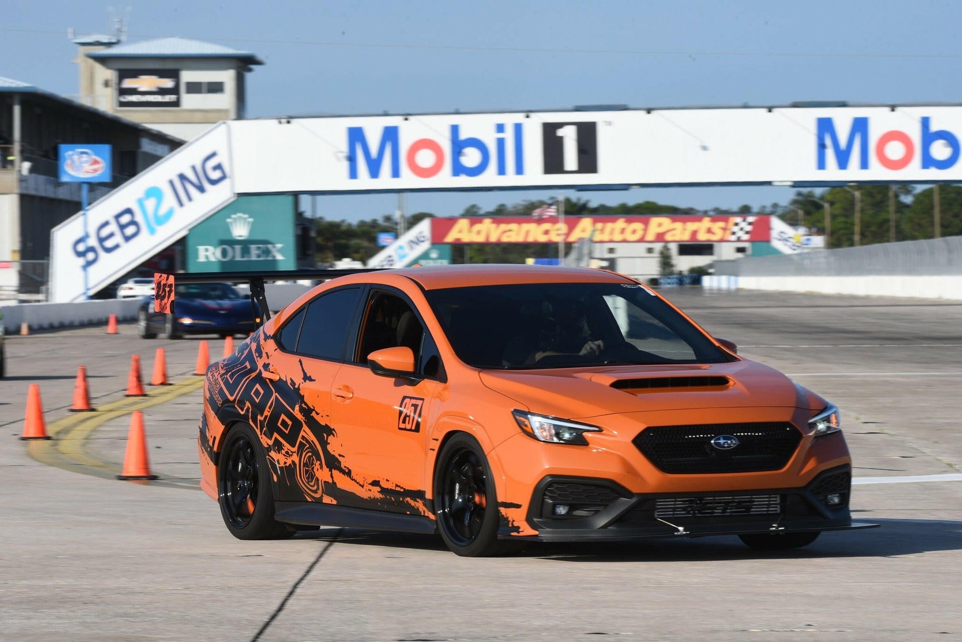 A Subaru Impreza STI Big Wang kit '2022 - current from 9livesracing, featuring an orange and black design with the number 77 on the side and precise downforce adjustments, speeds along the track at Sebring International Raceway. Various sponsor banners, including Mobil 1 and Advance Auto Parts, are visible in the background. Orange safety cones line the edges of the track.