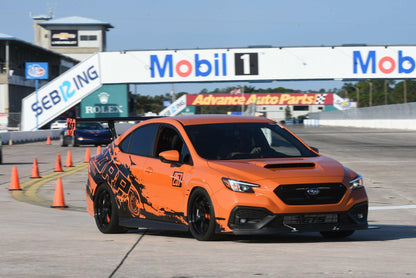 A Subaru Impreza STI Big Wang kit '2022 - current from 9livesracing, featuring an orange and black design with the number 77 on the side and precise downforce adjustments, speeds along the track at Sebring International Raceway. Various sponsor banners, including Mobil 1 and Advance Auto Parts, are visible in the background. Orange safety cones line the edges of the track.