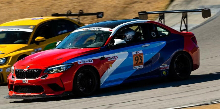 A race car with a red, blue, and black color scheme, marked with the number 93 and equipped with a 9livesracing 3-Series Big Wang kit ’14-18 F80 / F82 / F83 rear wing for optimal downforce, speeds around the track. A yellow race car is visible behind it, also racing on the track. The background shows a section of the raceway.