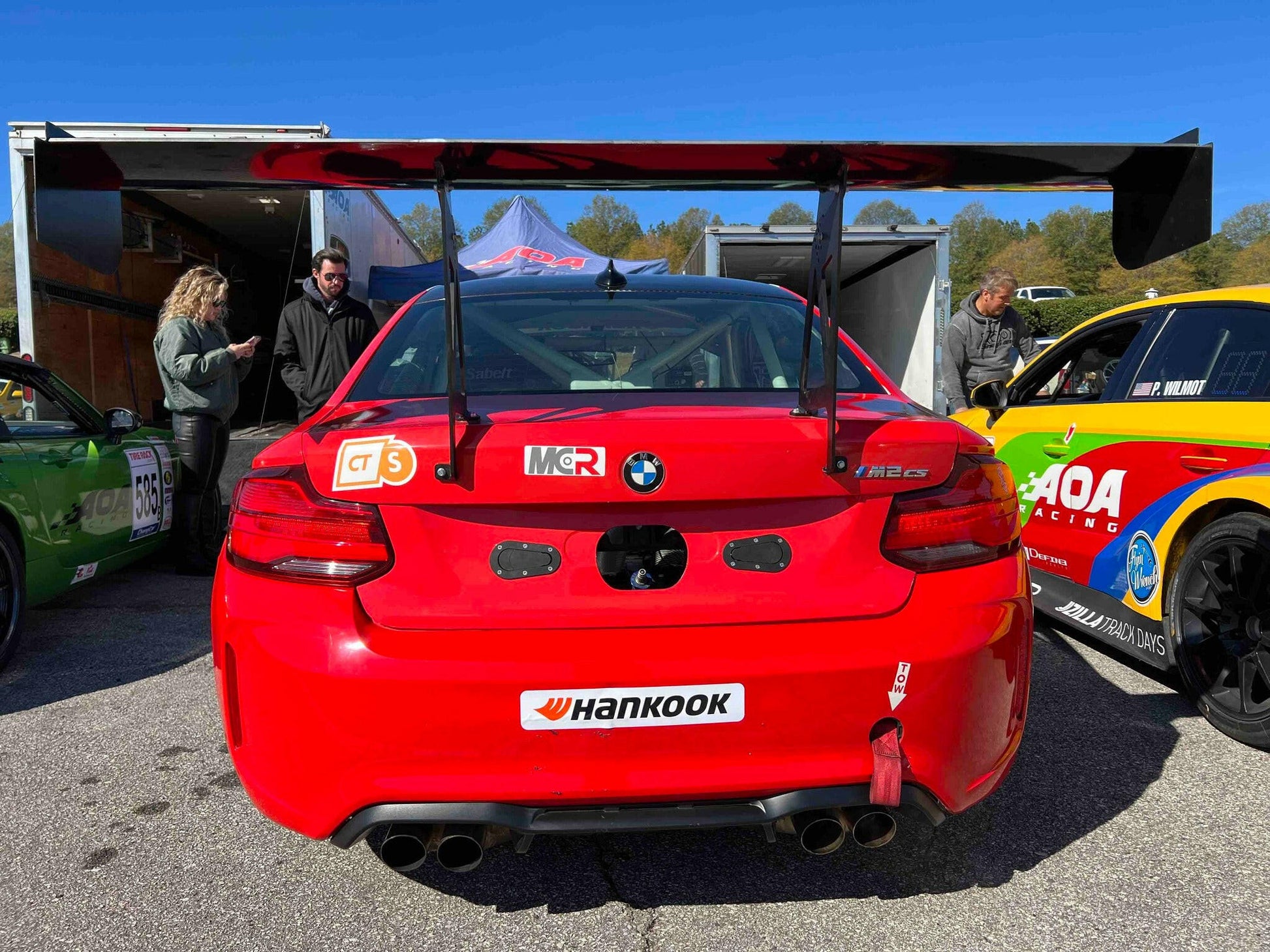A red race car featuring the 3-Series Big Wang kit ’14-18 F80 / F82 / F83 from 9livesracing is parked in a pit area. The car boasts several sponsorship stickers, including one from Hankook. People stand around the vehicle, appreciating its extraordinary downforce capabilities, while other race cars can be seen nearby.