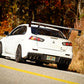 A white Mitsubishi Lancer Evolution, equipped with the Evolution 10 Big Wang kit from 9livesracing, gracefully navigates a winding road flanked by autumn-colored trees. The car boasts a prominent rear spoiler and aluminum wings, along with black rims and tinted windows, capturing the motion and vibrant fall foliage beautifully.