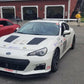 A white Subaru sports car, enhanced with a GT86 Big Wang Kit '12-'21 FRS / BRZ / 86 from 9livesracing, adorned with various decals and modifications including extruded aluminum wings for an improved lift-to-drag ratio, is parked in a lot among other cars, with a red one to the left and a yellow one to the right. Several people are standing around, and a red building with open garages is visible in the background.