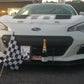 Front view of a white race car with a Subaru logo and the word "Hoosier" on the hood, proudly displaying a first-place plaque, a checkered flag, and a bottle of champagne on the front bumper. Equipped with the GT86 Big Wang Kit '12-'21 FRS / BRZ / 86 from 9livesracing for exceptional performance featuring extruded aluminum wings, it is parked alongside yellow and red cars in the lot.