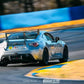 A sleek silver race car, labeled with the number 905, speeds on a race track. The car, featuring green and white livery and visible sponsor logos, boasts the 9livesracing GT86 Big Wang Kit for '12-'21 FRS / BRZ / 86 models with extruded aluminum wings for optimal lift-to-drag ratio. Trees and a blue barrier line the track in the background. Shot by Camden Thrasher for Yummi AER.
