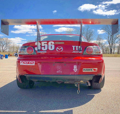 A red Mazda RX-8 race car with the number 356 and a large RX8 Big Wang Kit '03-12 SE3P aluminum wing by 9livesracing parked on an asphalt lot. The car features stickers from sponsors like NASA, Hawk, and Domo. Trees and a blue sky with scattered clouds are visible in the background.