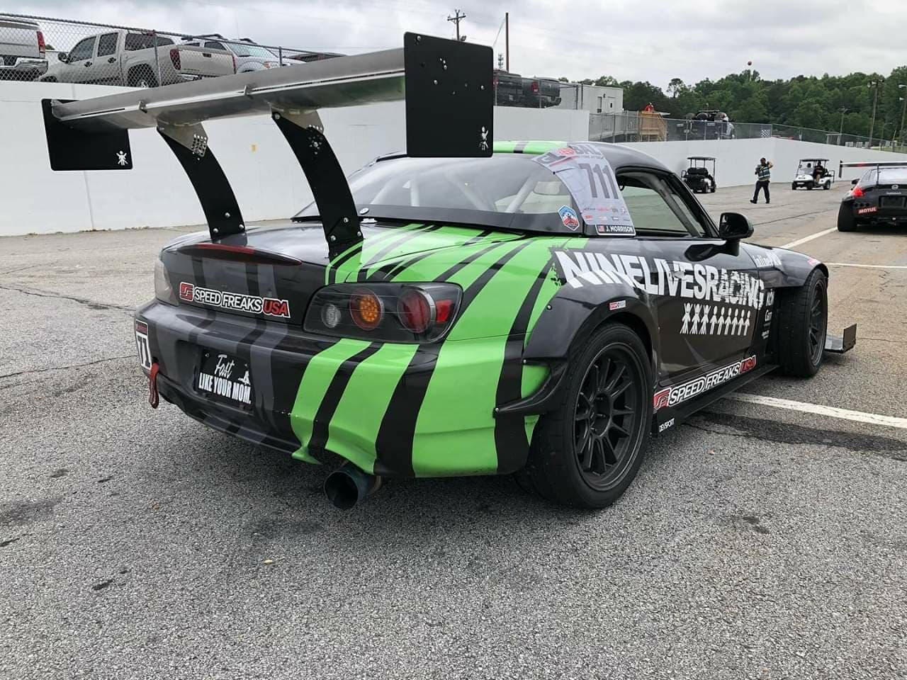 A race car with a prominent rear wing and a green-and-black striped design is parked in the pit area. The number "711" is displayed on the side window. The car features various decals, including "9livesracing" on the rear fender and "Speed Freaks USA" on the bumper, enhancing its downforce with sleek end plates from The Big Wáng!
