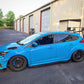 A modified blue hatchback featuring the Focus/RS Big Wang Kit '11-18 3rd Gen by 9livesracing, with a prominent aluminum rear wing and black wheels, is parked in front of a row of industrial garages. The car boasts additional aerodynamic parts, ensuring maximum down-force, and a lowered stance, indicating it is likely customized for performance or racing.