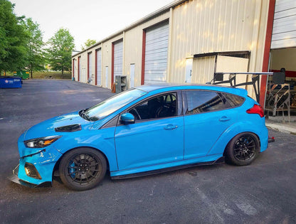 A modified blue hatchback featuring the Focus/RS Big Wang Kit '11-18 3rd Gen by 9livesracing, with a prominent aluminum rear wing and black wheels, is parked in front of a row of industrial garages. The car boasts additional aerodynamic parts, ensuring maximum down-force, and a lowered stance, indicating it is likely customized for performance or racing.
