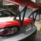 Close-up of the rear end of a Mazda Miata with a large custom Miata Big Wang Kit '06-15 NC *V1* spoiler from 9livesracing featuring a racing logo. The car has a red and white paint job and is parked in front of a stone-faced building. The license plate area displays an SCCA sticker.