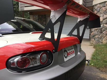 Close-up of the rear end of a Mazda Miata with a large custom Miata Big Wang Kit '06-15 NC *V1* spoiler from 9livesracing featuring a racing logo. The car has a red and white paint job and is parked in front of a stone-faced building. The license plate area displays an SCCA sticker.