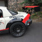 A close-up of the rear portion of a white Mazda race car with black and red accents. It features a large 9livesracing Miata Big Wang Kit '06-15 NC *V1* aluminum wing, black fender flares, and black multi-spoke wheels. The background shows a stone wall and greenery. The name "ROBBINS" appears near the roof.