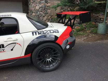 A close-up of the rear portion of a white Mazda race car with black and red accents. It features a large 9livesracing Miata Big Wang Kit '06-15 NC *V1* aluminum wing, black fender flares, and black multi-spoke wheels. The background shows a stone wall and greenery. The name "ROBBINS" appears near the roof.