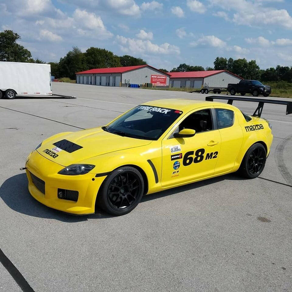 A yellow Mazda RX-8 race car with black wheels and the number 68M2 on the door is parked on a track. The car features various sponsor stickers, an aluminum 9livesracing RX8 Big Wang Kit '03-12 SE3P rear spoiler, and a vented hood. A white trailer and a blue sky with scattered clouds are visible in the background.