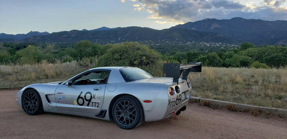 A silver racing car, featuring the 9livesracing Corvette Big Wang Chassis Mount ’97-13 C5/6 rear wing crafted from extruded aluminum for an optimal lift-to-drag ratio, is parked on a dirt path. The vehicle proudly displays racing decals such as "69x," "TTA," and "TT2." In the background, lush trees and a mountainous landscape stand against a cloudy sky during sunrise or sunset.
