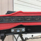 Close-up of the 9livesracing C10 1967-72 Truck Spoiler Kit, featuring a red component with a black adjustable ruler made from durable 6061 aluminum, all placed atop a metal stand. The background shows a corrugated metal wall and a closed garage door. Bolts and chains are attached to the lower section of the kit.