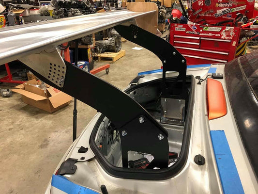 A race car with its rear trunk open, revealing a large custom Miata Big Chassis mount Wang Kit 2016+ ND from 9livesracing, mounted on black metal brackets. The car is in a workshop with various tools, parts, and equipment visible in the background, including red toolboxes and car parts on shelves, all engineered to optimize down-force.