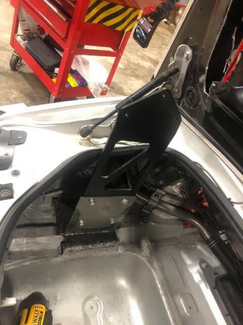 A partially disassembled car showcasing the Miata Big Chassis mount Wang Kit 2016+ ND by 9livesracing, with its black bracket and mounting hardware installed near the rear section, exhibits impressive down-force potential. In the background, a red toolbox adorned with yellow striped details is visible while various tools and components are scattered around the vehicle's interior.