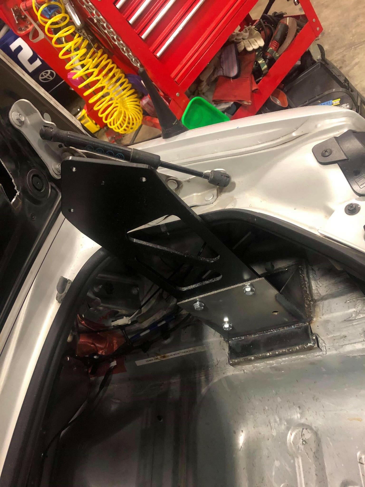 A close-up view of the interior of a vehicle trunk shows a metal mounting bracket installation. The bracket is part of the Miata Big Chassis mount Wang Kit 2016+ ND by 9livesracing and is attached near the trunk edge, designed to support aluminum wings for added down-force. Various tools and equipment can be seen in the background with a red tool chest and a yellow coiled hose.
