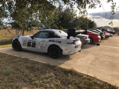 A white racing car featuring the number 62 and various sponsor logos is parked on a concrete lot next to other cars. It is equipped with the Miata Big Chassis mount Wang Kit 2016+ ND by 9livesracing, designed with sleek aluminum wings to maximize down-force. The scene unfolds under a partly cloudy sky, with trees and a fenced area in the background.