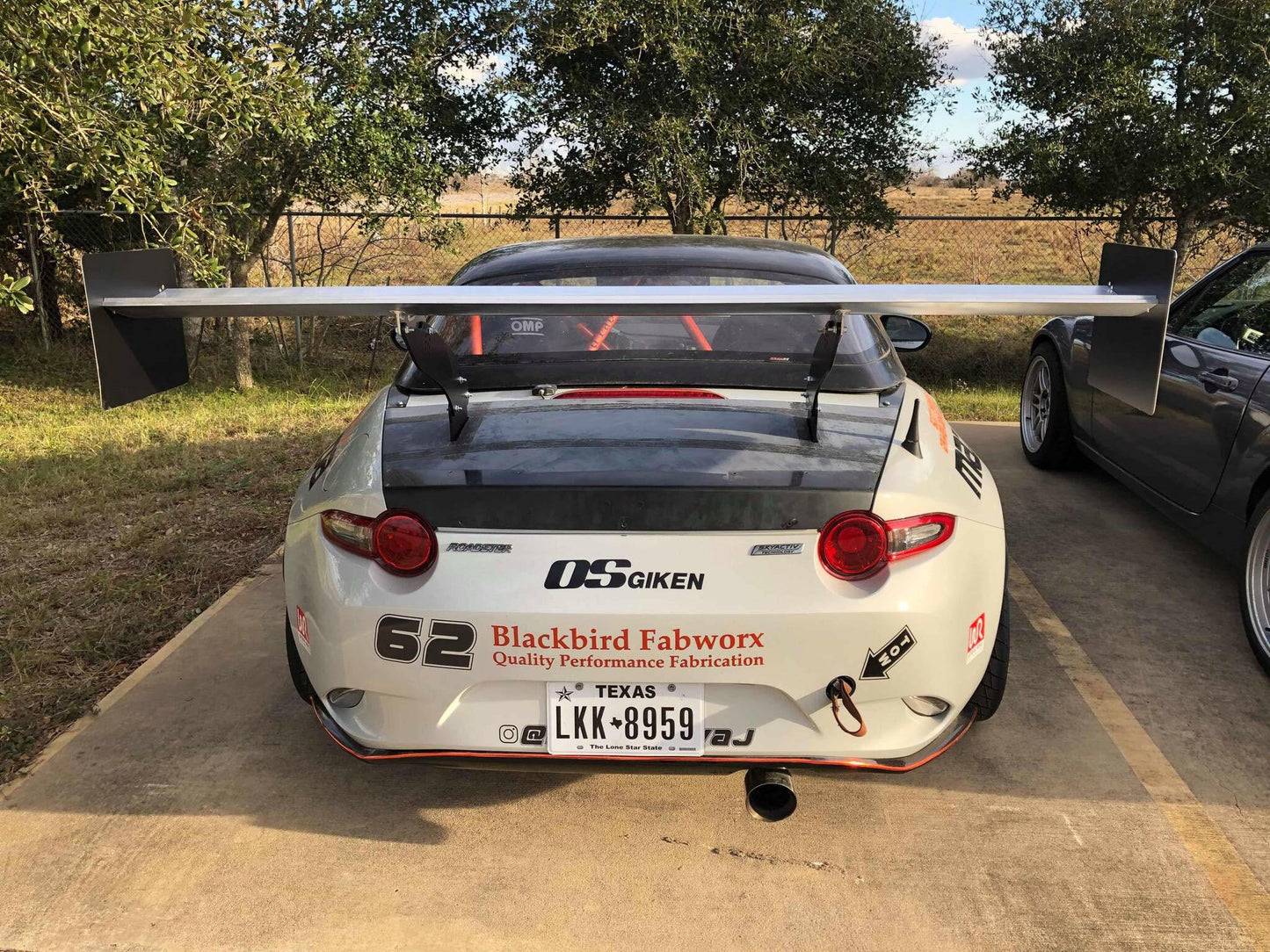 A modified white sports car displaying decals such as "OS Giken," "Blackbird Fabworx," and the number "62" boasts a Texas license plate reading "LKK-8959." The vehicle is equipped with a 9livesracing Miata Big Chassis mount Wang Kit 2016+ ND, providing substantial aluminum wings for enhanced down-force. It is parked next to another vehicle, with trees and a fence visible in the background.