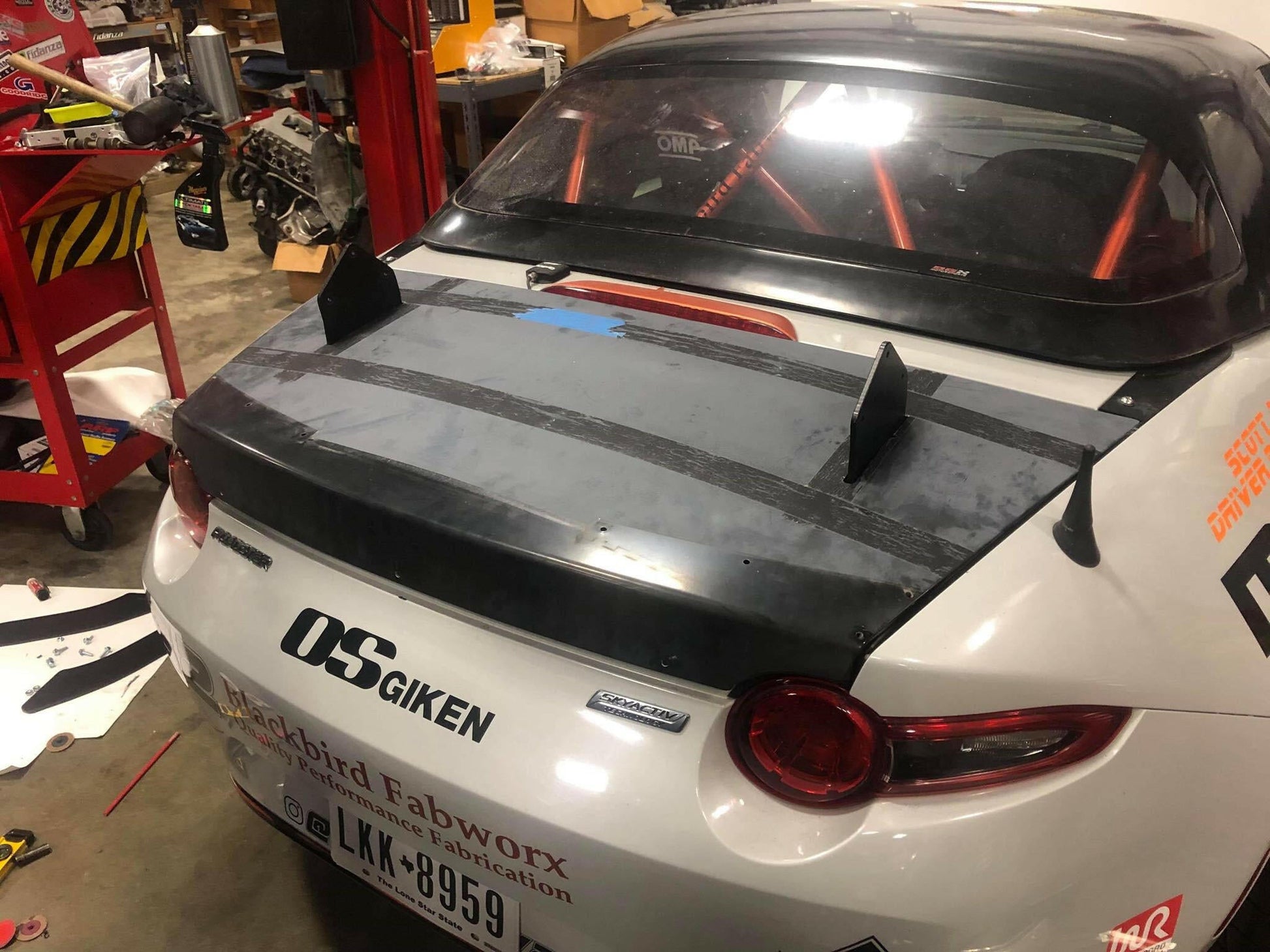 A close-up of the rear of a race car inside a garage reveals the car featuring a Miata Big Chassis mount Wang Kit 2016+ ND, by 9livesracing. The matte black spoiler with two vertical stabilizers, often referred to as a Big Wang for its imposing presence, stands out. Inside the car, a roll cage is visible. The license plate reads "LKK-8959," and several sponsorship decals adorn the exterior.