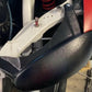 A close-up view of the Miata Headlight Blanks NB1-Only, a black, textured metal component from Nine Lives Racing, reveals its attachment to a white frame with screws. A red coiled spring is visible in the background, along with other metallic elements and partially visible robust plastic components that contribute to its durability.