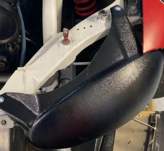 A close-up view of the Miata Headlight Blanks NB1-Only, a black, textured metal component from Nine Lives Racing, reveals its attachment to a white frame with screws. A red coiled spring is visible in the background, along with other metallic elements and partially visible robust plastic components that contribute to its durability.
