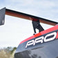 Close-up of the rear wing, specifically the Mustang Big Wang Kit '10-14 S197 II by 9livesracing, featuring an extruded aluminum performance airfoil, on a black and red sports car with the word "PROA" partially visible on the side, set against a blurred background of trees and a cloudy sky.