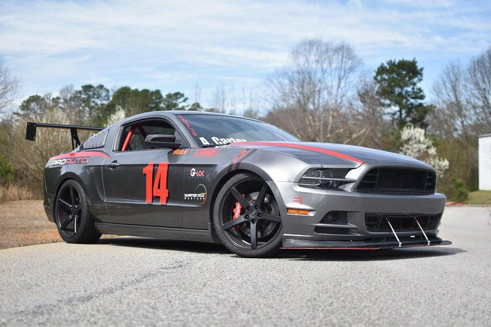 A gray Mustang Big Wang Kit '10-14 S197 II, proudly displaying the number 14 in red on its side, is parked on a road. This car from 9livesracing boasts various decals, black rims, and a prominent front splitter with an extruded aluminum wing designed for optimal downforce adjustment. In the background, trees and a patch of blue sky with some clouds can be seen.