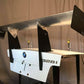 A desk constructed from the rear end of a BMW 328i car, outfitted with the 9livesracing 3-Series Big Wang Kit '90-00 E36, showcasing a prominent metal racing spoiler supported by black brackets. The car's license plate space, logo, and model designation remain clearly visible. The desk surface is white and set against a backdrop of a plain curtain.