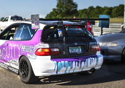 A modified Honda hatchback, equipped with a 9livesracing Civic Hatch EG Wang kit for the 92-95 model, boasts impressive down-force thanks to its Big Wang rear wing and vibrant graphics on the bumper. It is parked on a paved area near other cars. The custom license plate reads "OMGIT4K." In the background, greenery and another vehicle add to the scene.